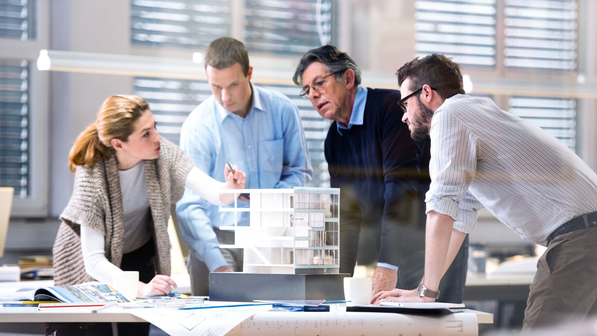 Architect explaining project plan to client in office with the help of an architectural model. Project manager and co-worker standing beside him.