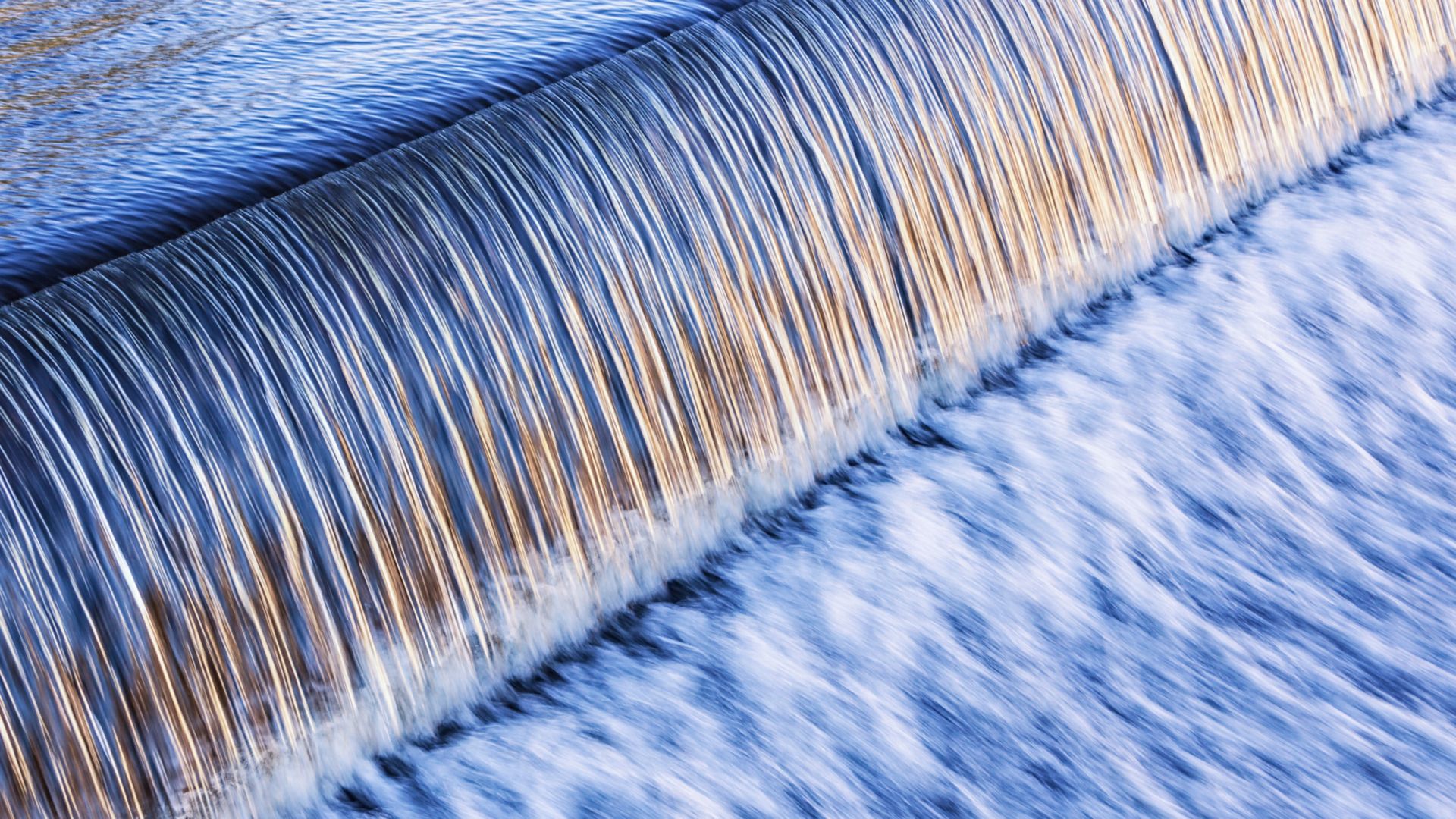 Rushing water flows over a man-made waterfall at the edge of an early evening Lake Placid, New York municipal river dam. Letterbox format composition.