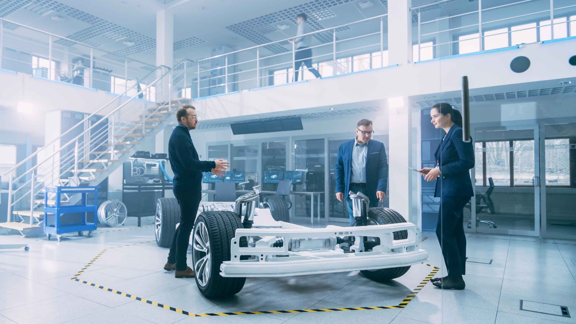 Automotive Design Engineers Talking while Working on Electric Car Chassis Prototype. In Innovation Laboratory Facility Concept Vehicle Frame Includes Wheels, Suspension, Engine and Battery.