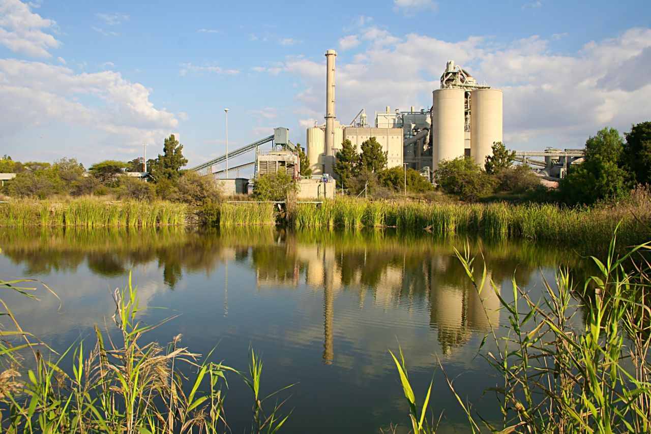 Cement production plant next to lake with reflection, grass, trees, blue sky
