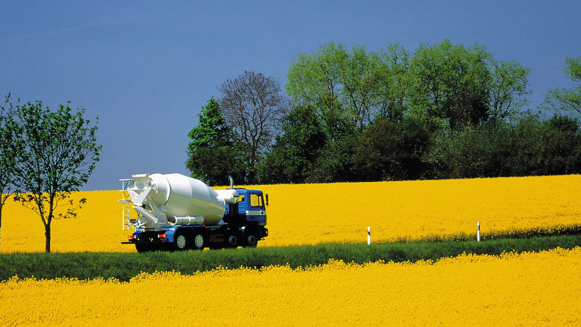 Ready-mix concrete mixing truck driving through yellow field with trees and blue sky