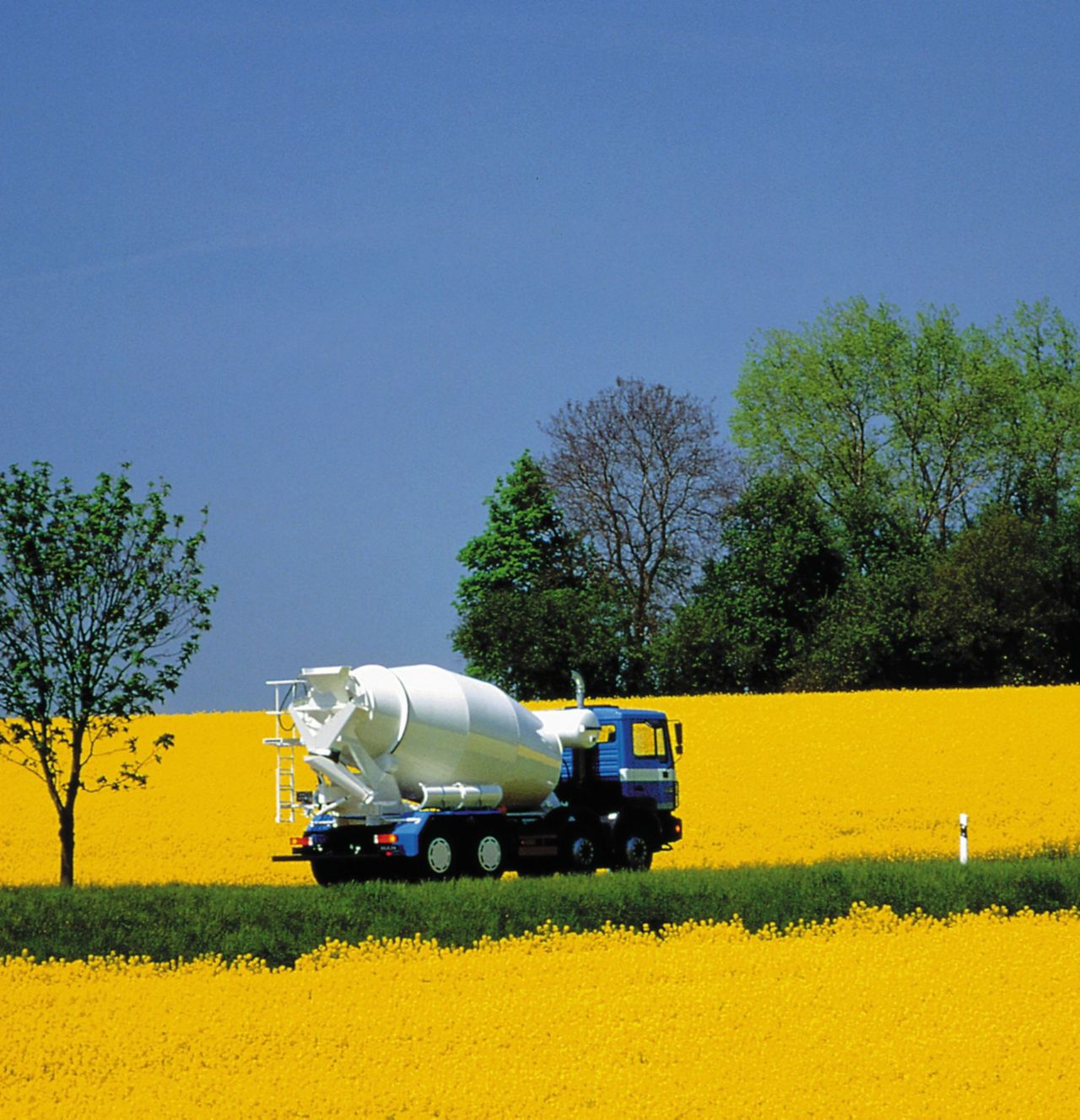 Ready-mix concrete mixing truck driving through yellow field with trees and blue sky