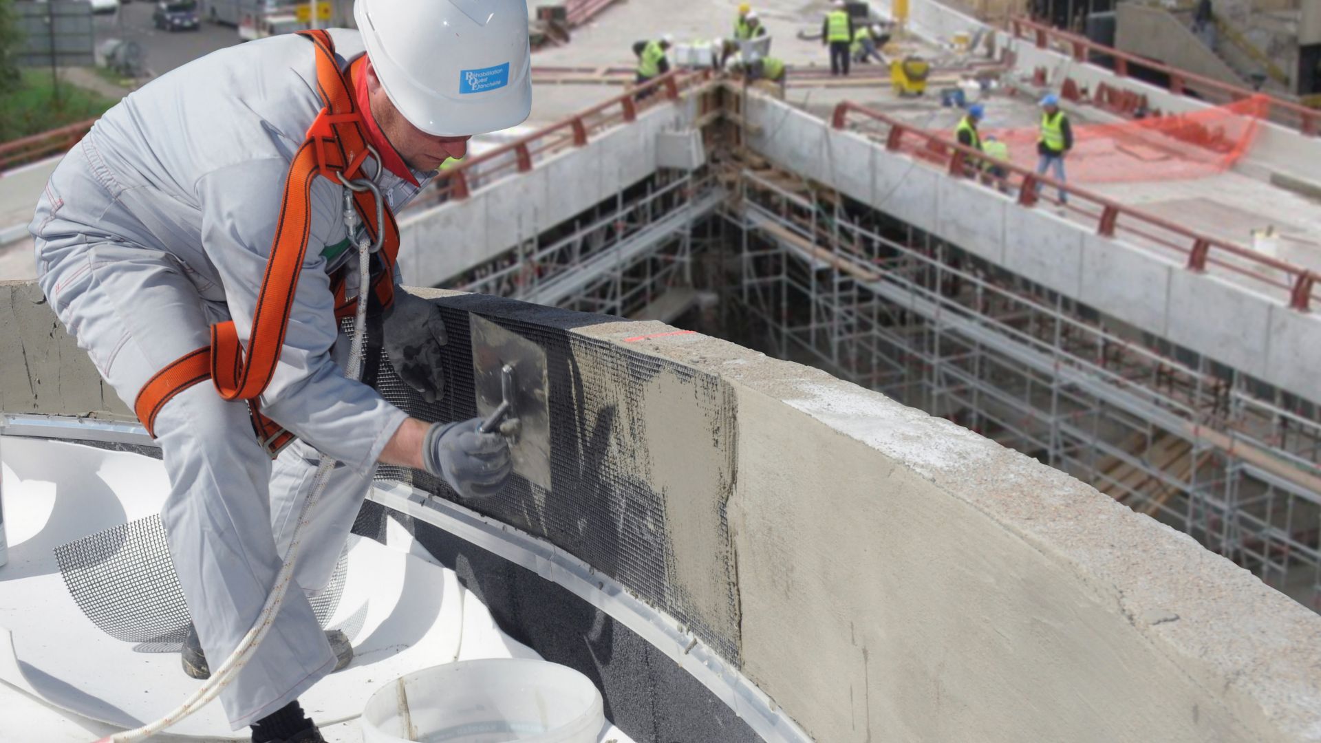 Man applying mortar over carbon grid reinforcement on FRCM wall with bridge in background