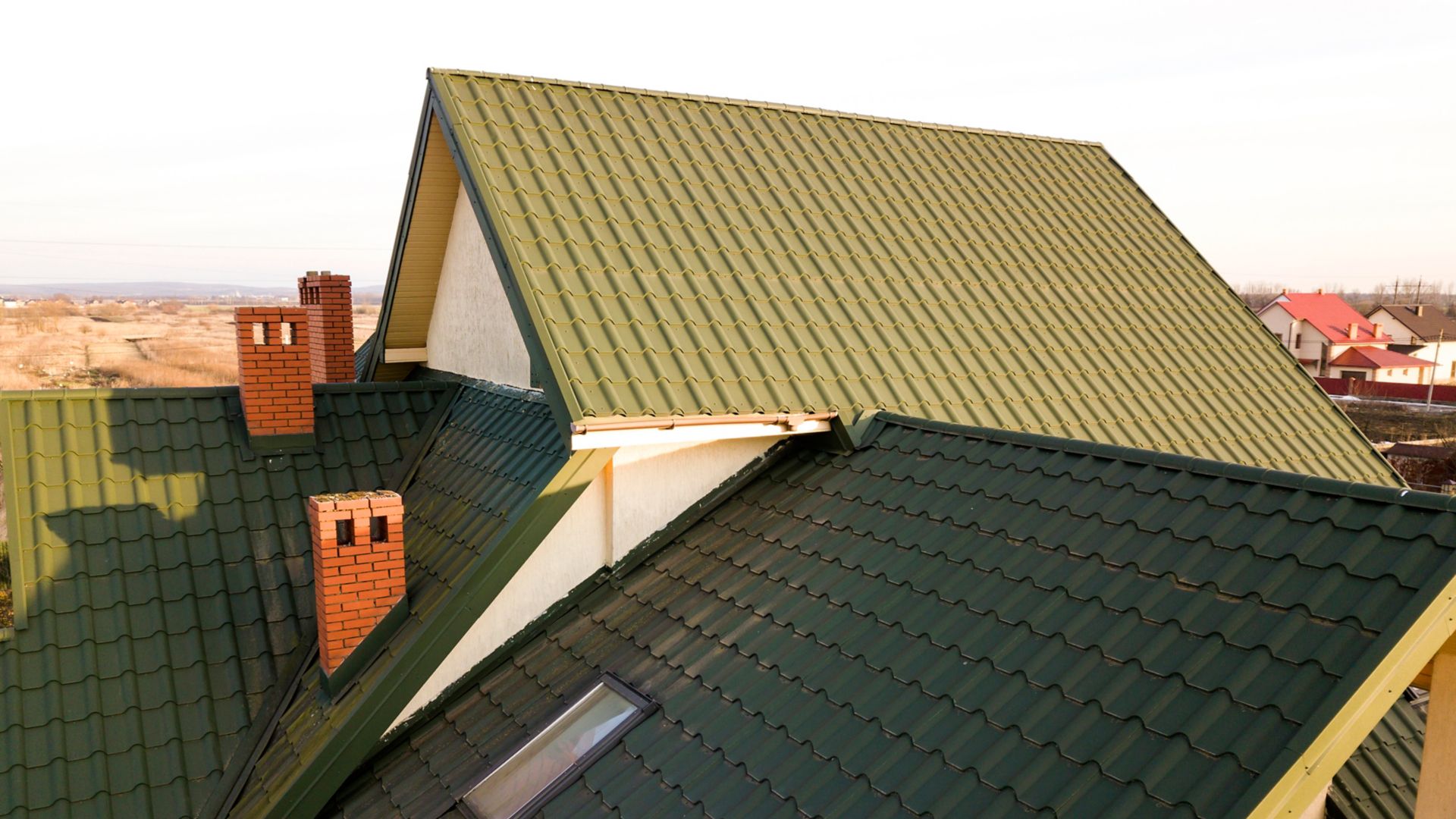 Green metal shingled house roof with attic plastic window and brick chimney.