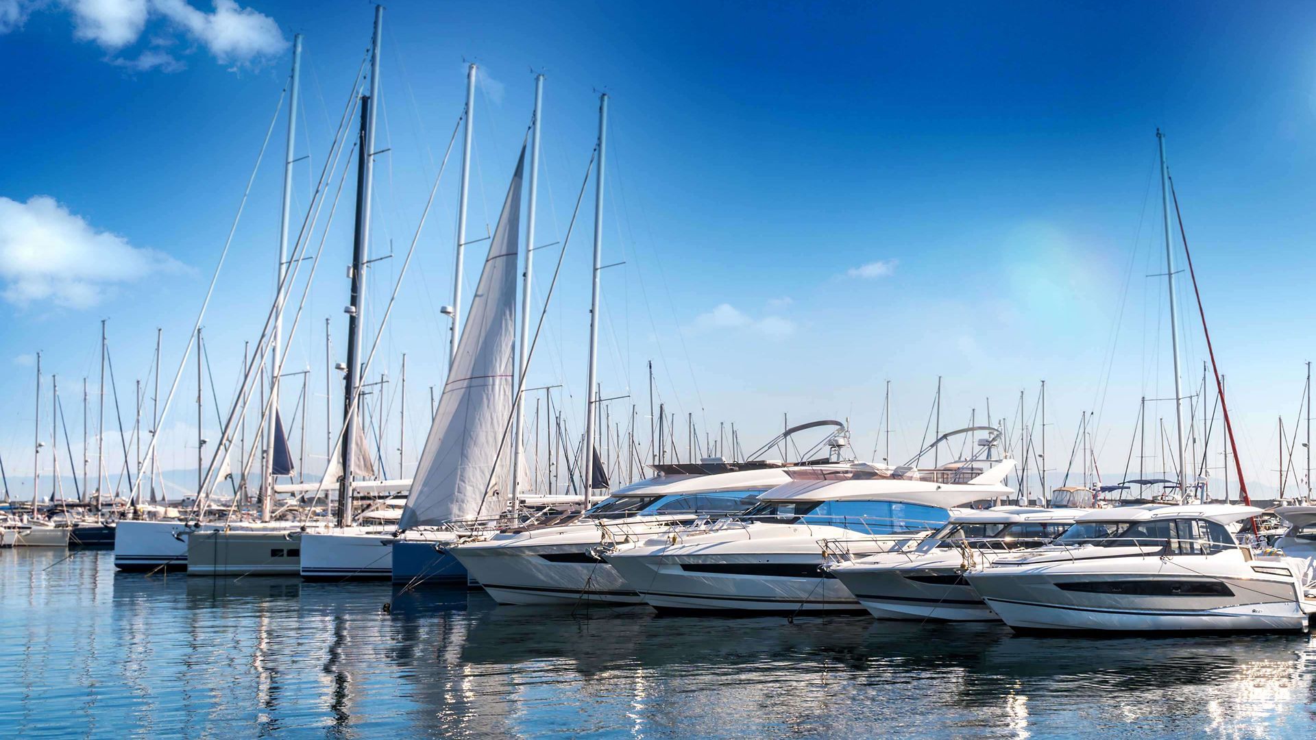 Marina with yachts and sail boats lined up in blue water.  Blue skies with clouds.