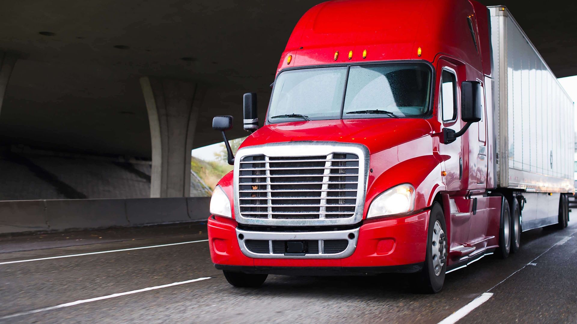 red semi-truck with trailer riding on freeway under bridge; heavy truck