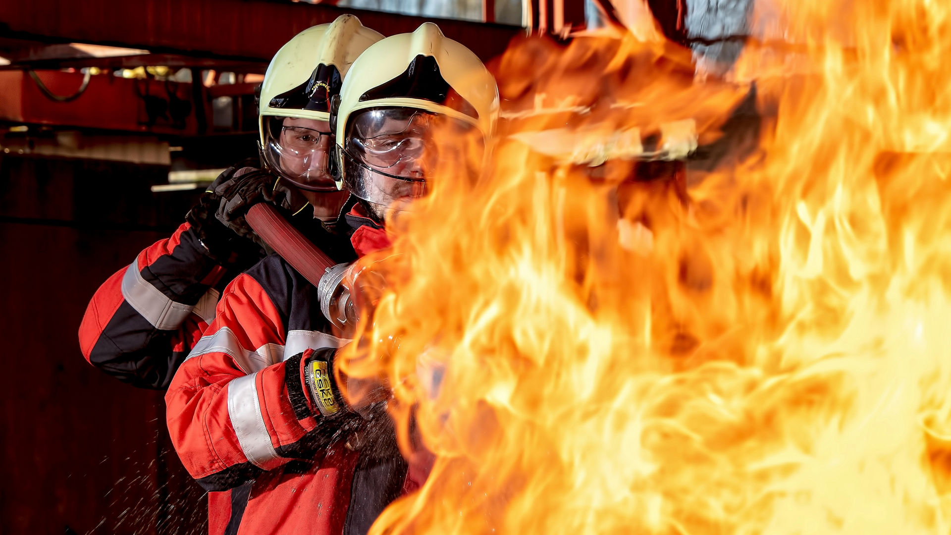 2 firefighters carying a waterhose on their shoulders, extinguishing a fire