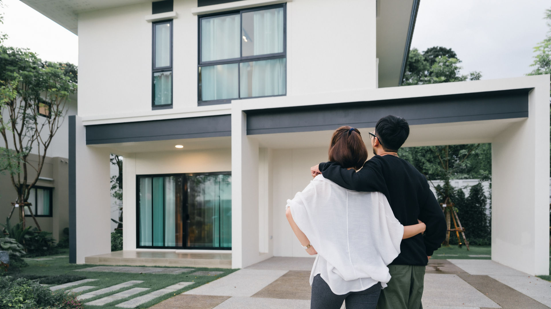 Asian dark haired couple embracing in front of white facade new home